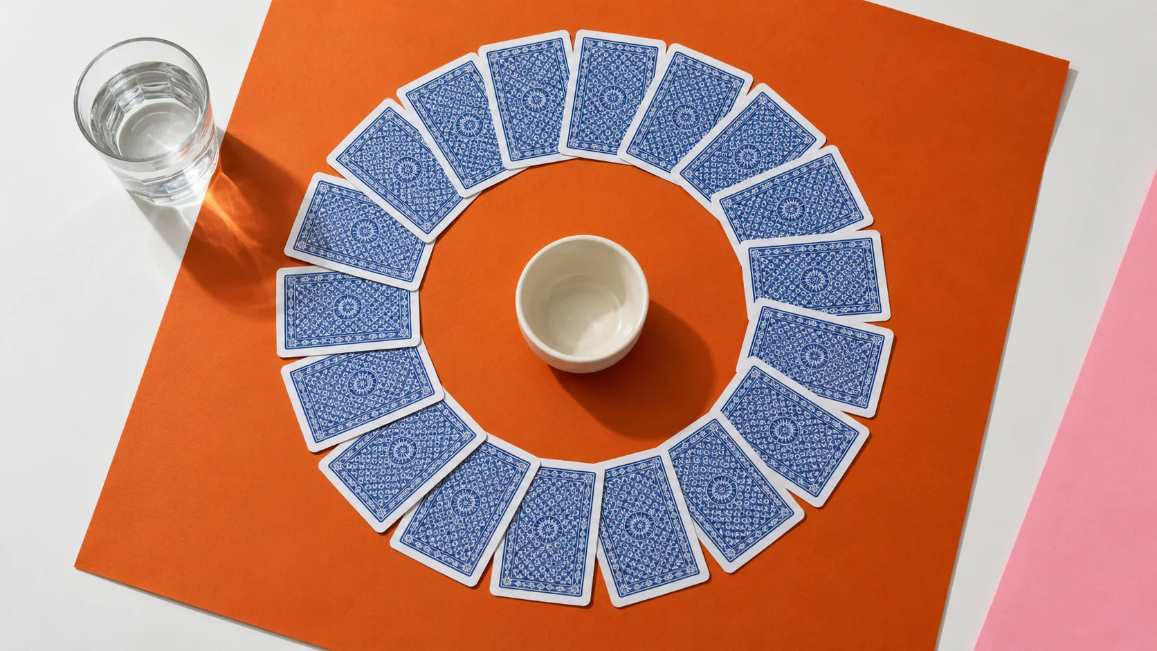 A ring of playing cards around a small cup on a bold orange and cream tabletop, with water nearby.