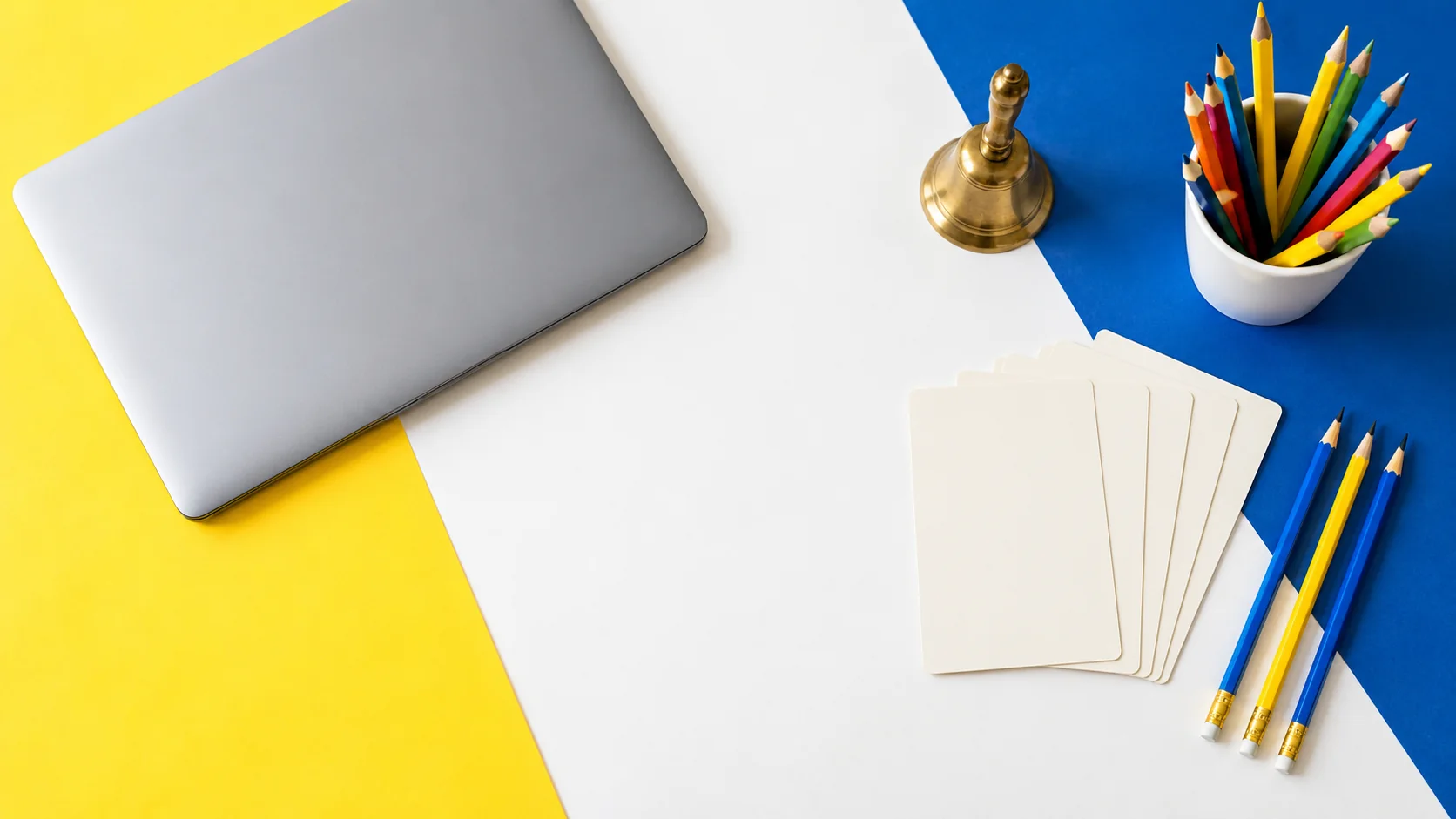 Colored pencils, a small classroom bell, blank vocabulary cards and a laptop on a yellow and blue desk.